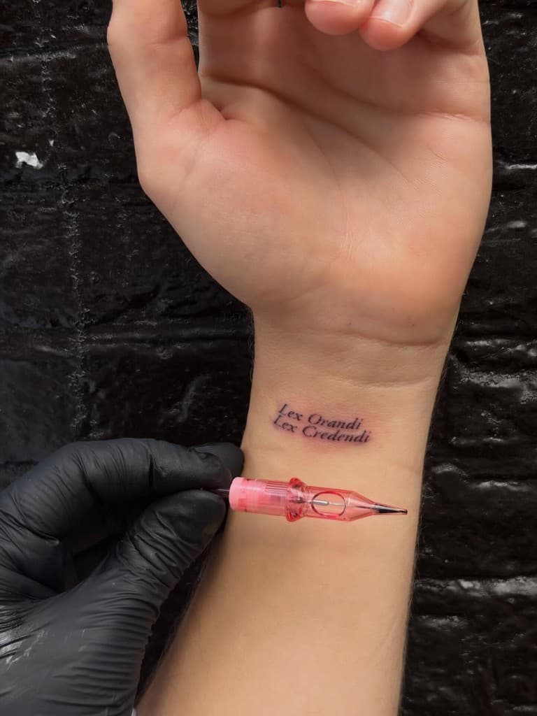 Fresh two-line italic script tattoo on an inner wrist reading Lex Orandi Lex Credendi, with a tattoo cartridge in frame.